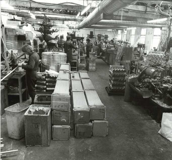 Greyscale photo of the interior of the manufacturing facilities at JDR Products LTD. There are pallets of bundled aluminium tubes and billets with several people seen working at machinery.